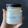 Load image into Gallery viewer, Hand holding a candle jar labeled 'Coastal Wind & Lavender' against a dark background