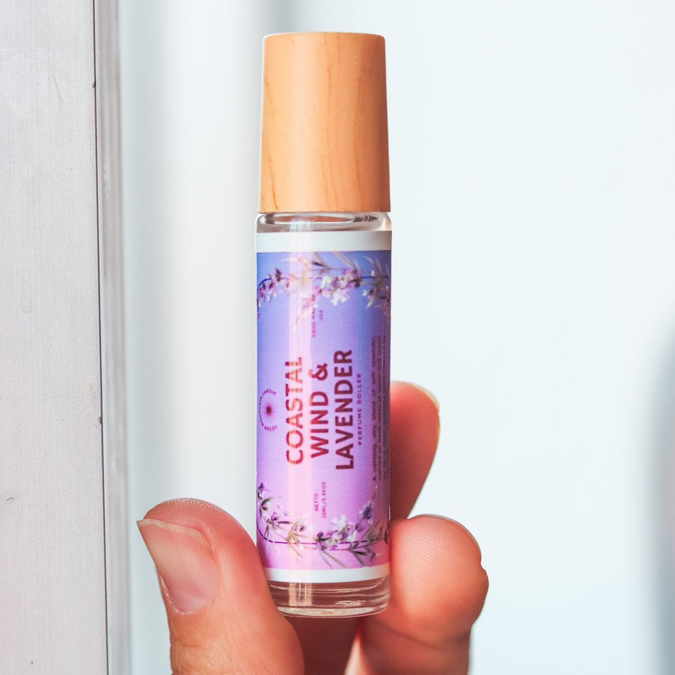 Hand holding a small bottle labeled 'Coastal Wind & Lavender' against a blurred background